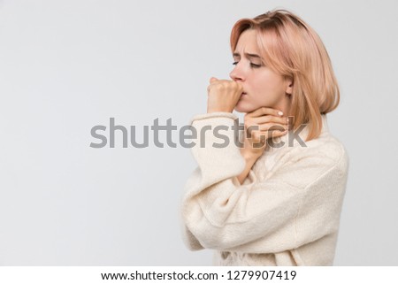 Studio portrait of young unhealthy coughing blonde woman in warm sweater/suffers from cold and flu, coughing a lot, sore throat, feeling sick, chest pain, medicine and healthcare concept. Royalty-Free Stock Photo #1279907419