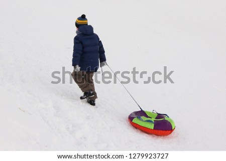 Child in blue jacket and hat pulls on the rope inflatable snowtubing in the mountain on winter day - side rear view