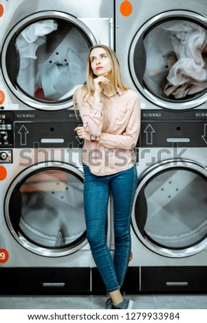 Young woman with boring emotions standing near the dryer machines waiting for clothes to be dried in the laundry
