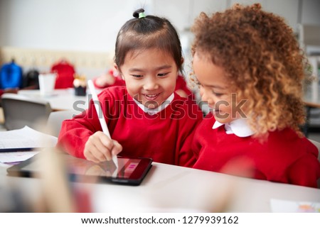 Close up of two kindergarten schoolgirls wearing school uniforms, sitting at a desk in a classroom using a tablet computer and stylus, looking at the screen and smiling Royalty-Free Stock Photo #1279939162