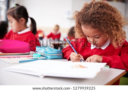 Mixed race schoolgirl wearing school uniform sitting at a desk in an infant school classroom drawing, close up Royalty-Free Stock Photo #1279939225