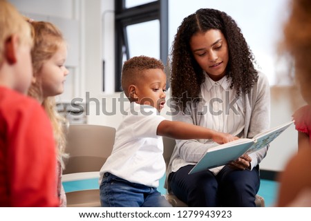 Infant school boy pointing in a book held by the female teacher, sitting with kids on chairs in the classroom, close up Royalty-Free Stock Photo #1279943923