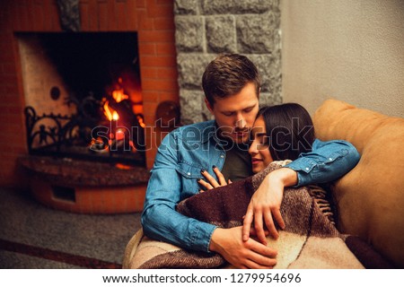 Lovely picture of young couple sleeping together. She leans to him on his chest. Model coveres with blanket. Guy embrace her. They sit near fireplace.