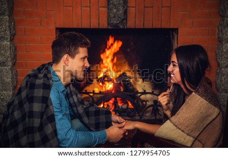 Cozy picture of lovely young man and woman look at each other and smile. They have blankets on shoulders. Young couple sit at fireplace with fire.