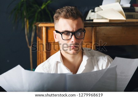 Handsome writer man wearing glasses and white shirt sitting on the floor near the table with typewriter and looking on the new chapter of his book.