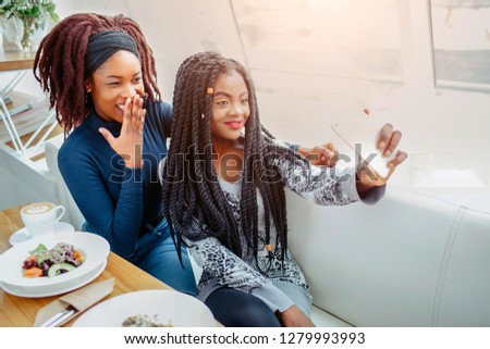 Cheerful nice young african women in cafe. They take selfie on phone camera. Models smile. They sit at table.