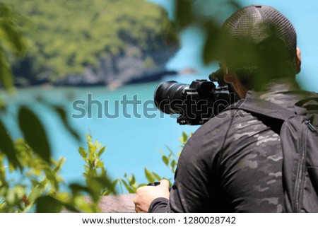 Photographer or videographer holding the video camera on a tripod on top of the mountain. Forest,ocean and blue sky  background.