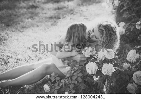 Mom kissing daughter on green grass on sunny summer day. Childhood and parenting. Family love and care. Woman with girl child at blossoming rose flowers. Mothers day concept.