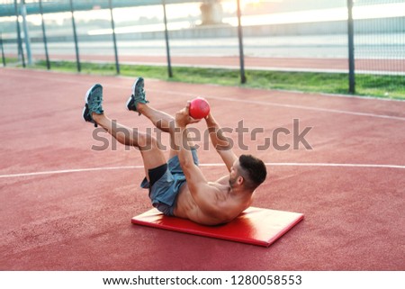 Dedicated shirtless muscular Caucasian man with short haircut sitting on the mat and doing exercises with kettlebell. Outdoor fitness concept.