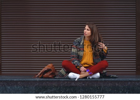Portrait of young smiling woman sitting crossed legs, posing in street of a European city. Braun copy space in background on the right