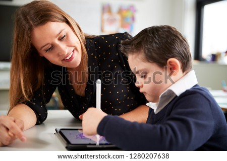 Close up of young female teacher sitting at desk with a Down syndrome schoolboy using a tablet computer in a primary school classroom, smiling, close up, side view Royalty-Free Stock Photo #1280207638