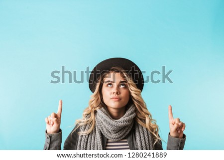 Pretty young girl wearing hat and a scarf standing isolated over blue background, pointing at copy space