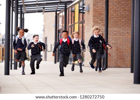 Primary school kids, wearing school uniforms and backpacks, running on a walkway outside their school building, front view Royalty-Free Stock Photo #1280272750