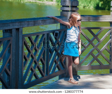 A little girl leaning against a railing of a deck along a pond at a park resting after taking pictures with her camera.