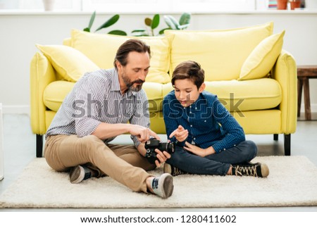 Calm attentive middle aged man sitting on the floor and showing his son a modern camera