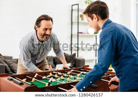 Emotional bearded man touching the control knobs and smiling while standing in front of his son in the table football game