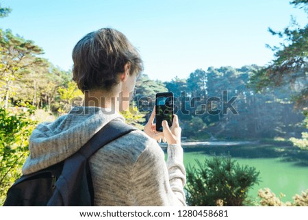 Back view close up young man with backpack filming video or taking photo pictures on his mobile cell phone during his adventure in the forest. Selective focus. Copy space.