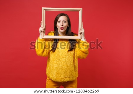 Portrait of amazed attractive young woman in yellow fur sweater holding picture frame isolated on bright red wall background in studio. People sincere emotions, lifestyle concept. Mock up copy space