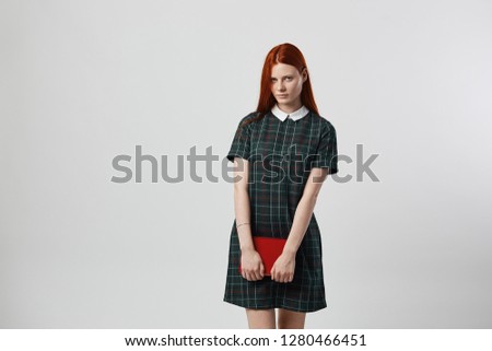 Charming redhead long-haired girl in a green checkered dress holds a book in her hands on the white background in the studio