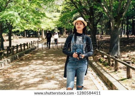 Asian chinese photographer woman holding camera standing outdoors under tree shadow in spring. young girl travel lens man wearing straw hat looking around in nara park japan. lady tourist in path.