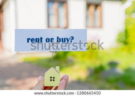 miniature house in hands with text rent or buy and question mark..Real estate concept. Business card with buy or rent message.Small model of house over country house.loans, Bank debt,
