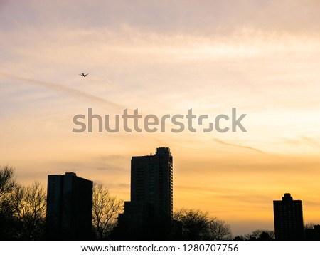 A commercial jet plane flies over residential condominium high rise buildings and trees along Lake Shore Drive and Lake Michigan in Chicago at sunset with beautiful orange cloudy sky beyond.