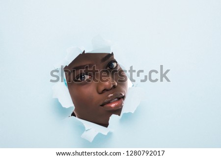 The face of a young beautiful african girl with a bright make-up and puffy lips peers into a hole in blue paper. Fashion, beauty, make-up, cosmetics, hairstyle, beauty salon.