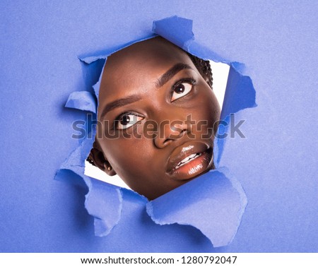 The face of a young african girl with a bright make-up and puffy purple lips peers into a hole in violet paper.