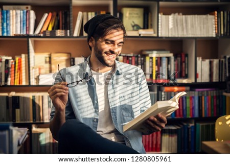 Cute smiling hipster boy sitting on a library floor and looking through some books.
