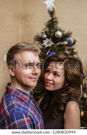A girl with long curly hair in a shiny festive top and a man with a blonde in a plaid shirt on the background of christmas tree