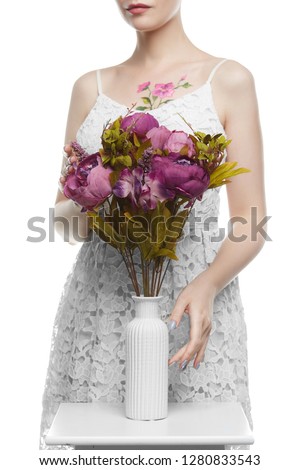 Shot of a lady with floral tattoo, wearing a white dress, posing on white background. The woman is touching bouquet of lavender peonies and a vase with fluted surface, which stands on small table.