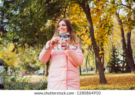 Joyful happy young woman in stylish pink coat and scarf on a background of yellow autumn park. Woman blowing soap bubbles outdoor. Conception of happy adult people who don't care to be like kids.