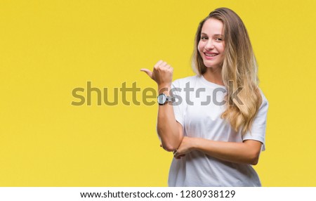 Young beautiful blonde woman wearing casual white t-shirt over isolated background smiling with happy face looking and pointing to the side with thumb up.