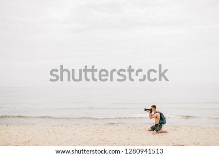 young man with shorts and a backpack behind him kneels on beach and takes pictures of anything. Photographer at sea with a professional camera shoots seascape. concept of recreation and tourism