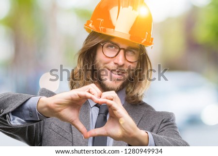 Young handsome architec man with long hair wearing safety helmet over isolated background smiling in love showing heart symbol and shape with hands. Romantic concept.