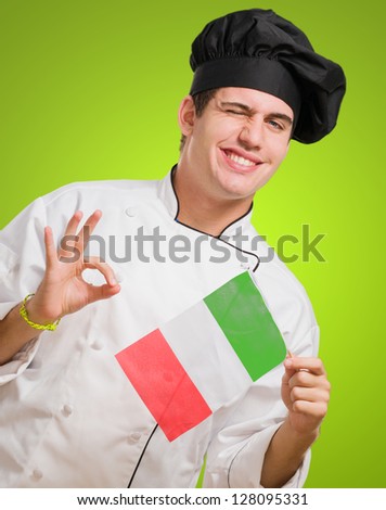 Portrait Of A Young Chef Holding Italian Flag And Winking against a green background