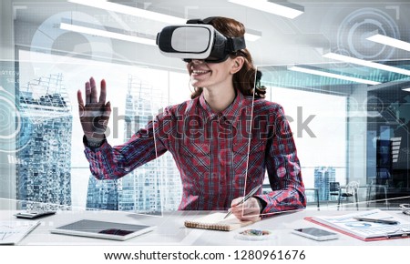 Portrait of beautiful and young woman in red checkered shirt using VR goggles and interracting with digital media interface while sitting inside bright office building. Virtual reality device