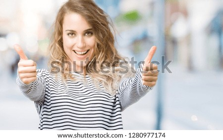 Beautiful young blonde woman wearing stripes sweater over isolated background approving doing positive gesture with hand, thumbs up smiling and happy for success. Looking at the camera, winner gesture