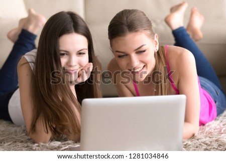 Portrait of two happy smiling young women friend lying on floor at home in front of laptop. Attractive ladies watching photo or video on computer, chatting in Internet, browsing news, shopping online