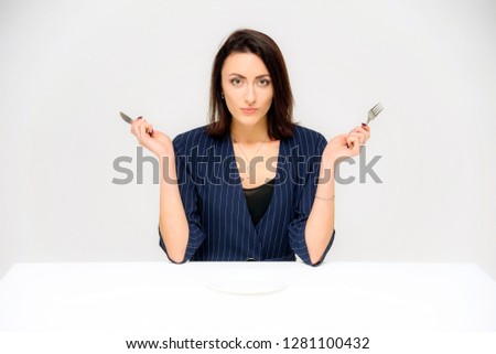 Concept portrait of a brunette girl, sitting on a white background in a business suit with a plate, fork, spoon. She sits right in front of the camera in various poses with different emotions.