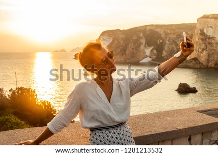 Young woman with smartphone taking selfies with sunset in front of the sea on Ponza island coast, on a wall with view of the ocean.