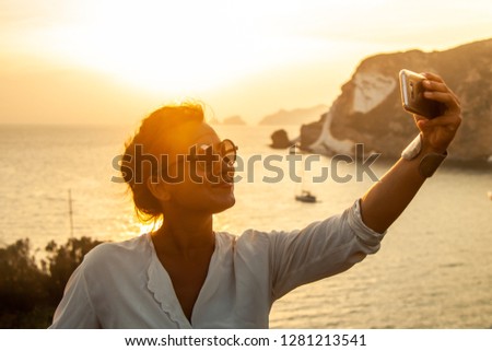 Young woman with smartphone taking selfies with sunset in front of the sea on Ponza island coast, on a wall with view of the ocean. 