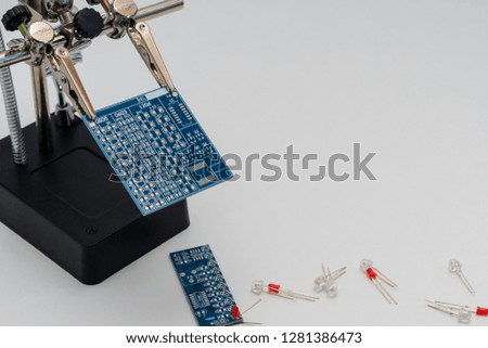 Blue circuit board in soldering helping hands (third hand or auxiliary clip tool) before technological soldering process with white and red  LEDs (Light-emitting diode) on the light background.