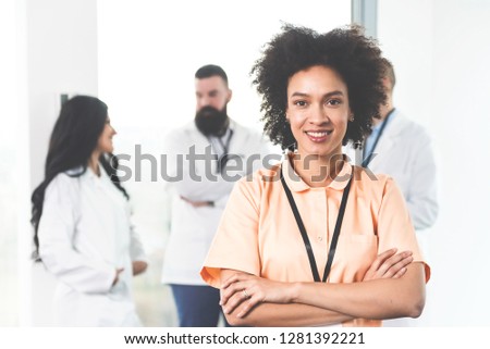 Closeup portrait of confident smart afro-americain female doctor posing with her arms crossed and looking at camera in her modern bright office, Doctors consulting in the background