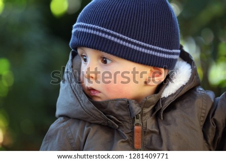 Close Up Portrait Of Cute Baby Boy Wearing A Blue Knit Winter Hat And Green Winter Parka