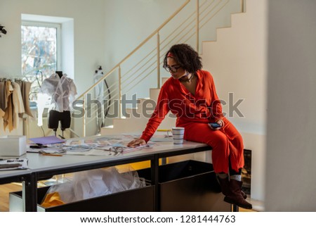 Cup of tea. Curly young woman with smartphone wearing long dress and observing photo materials on table surface