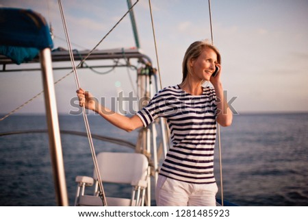 Mid-adult woman holding onto wire on boat as she looks out to view of sunset and talks on cell phone.