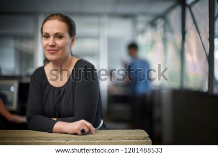 Businesswoman leans on a low, stone wall office partition and smiles as she poses for a portrait.