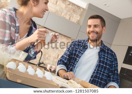 Husband and wife at home standing in kitchen together man rolling dough on board smiling cheerful looking at woman holding cutted heart shape dough