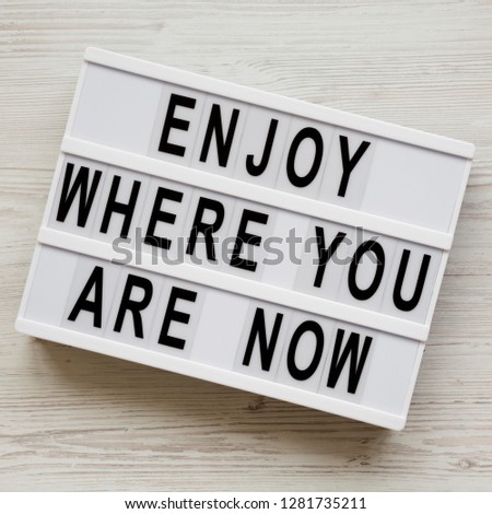 'Enjoy where you are now' words on modern board over white wooden surface, top view. Flat lay, from above.
